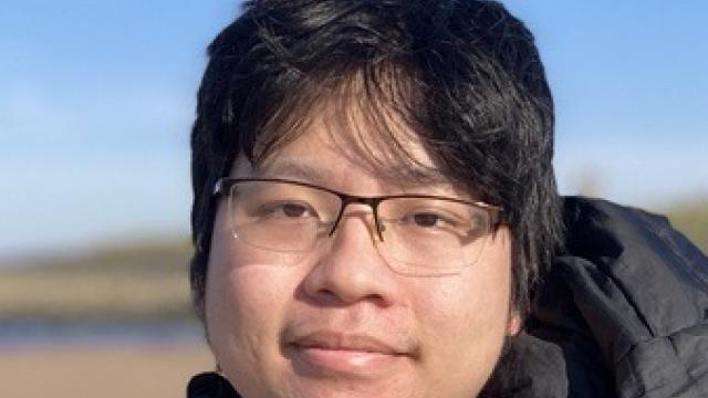 a close up of the face of a man with glasses, wearing a black puffer jacket on a beach